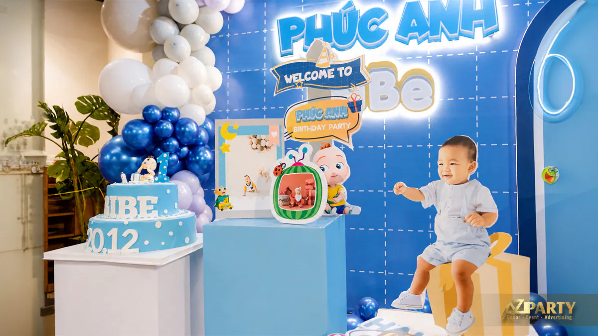 Cậu bé Phúc Anh đáng yêu ngồi trên mô hình hộp quà bên cạnh chiếc bánh kem màu xanh hai tầng
