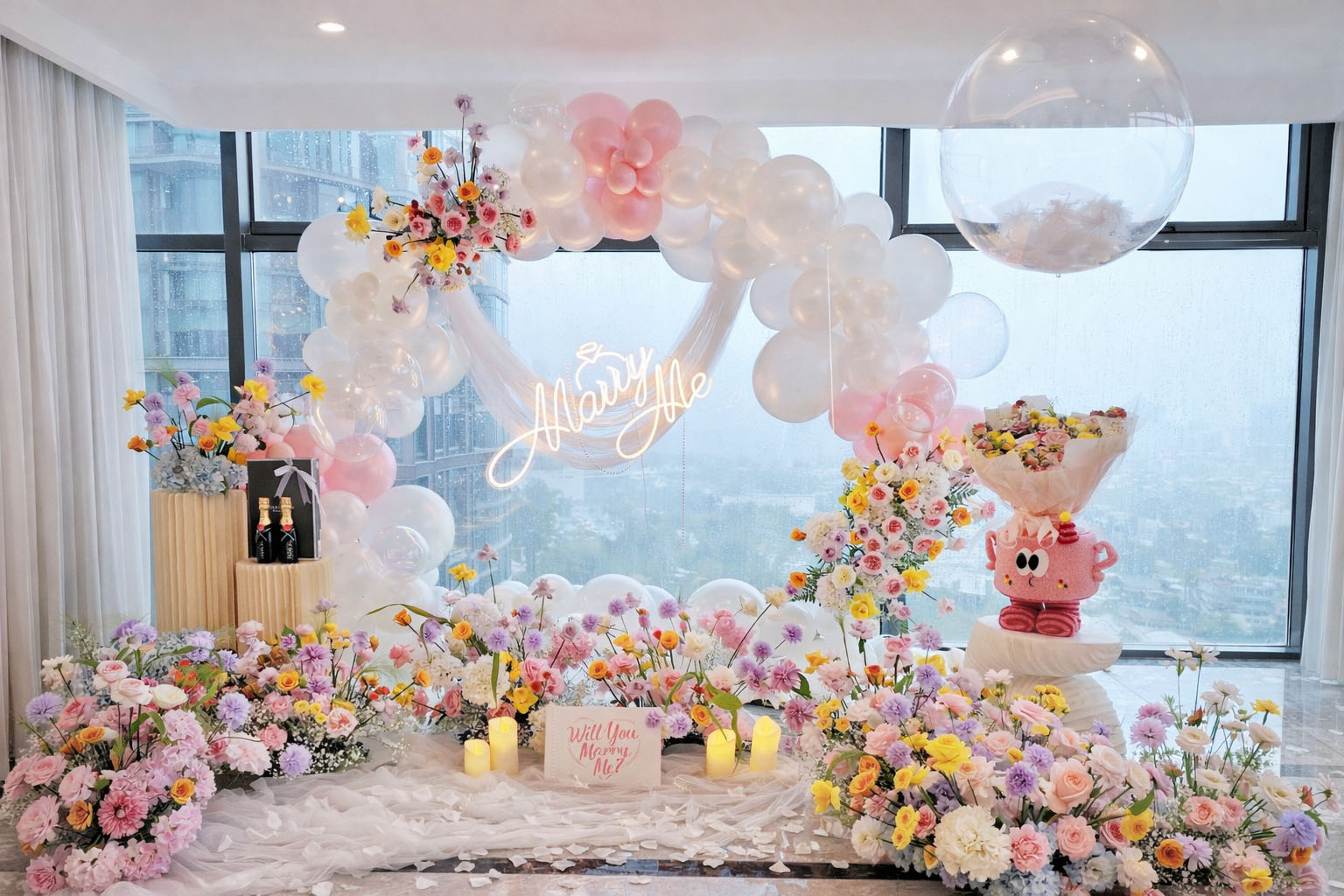 Không gian cầu hôn trong nhà với vòng bong bóng trắng hồng, hoa tươi và đèn chữ Marry Me lãng mạn.