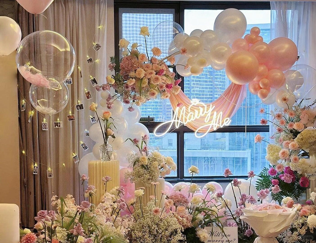 Trang trí cầu hôn trong nhà với vòng bong bóng trắng hồng, hoa tươi và đèn chữ Marry Me lãng mạn.