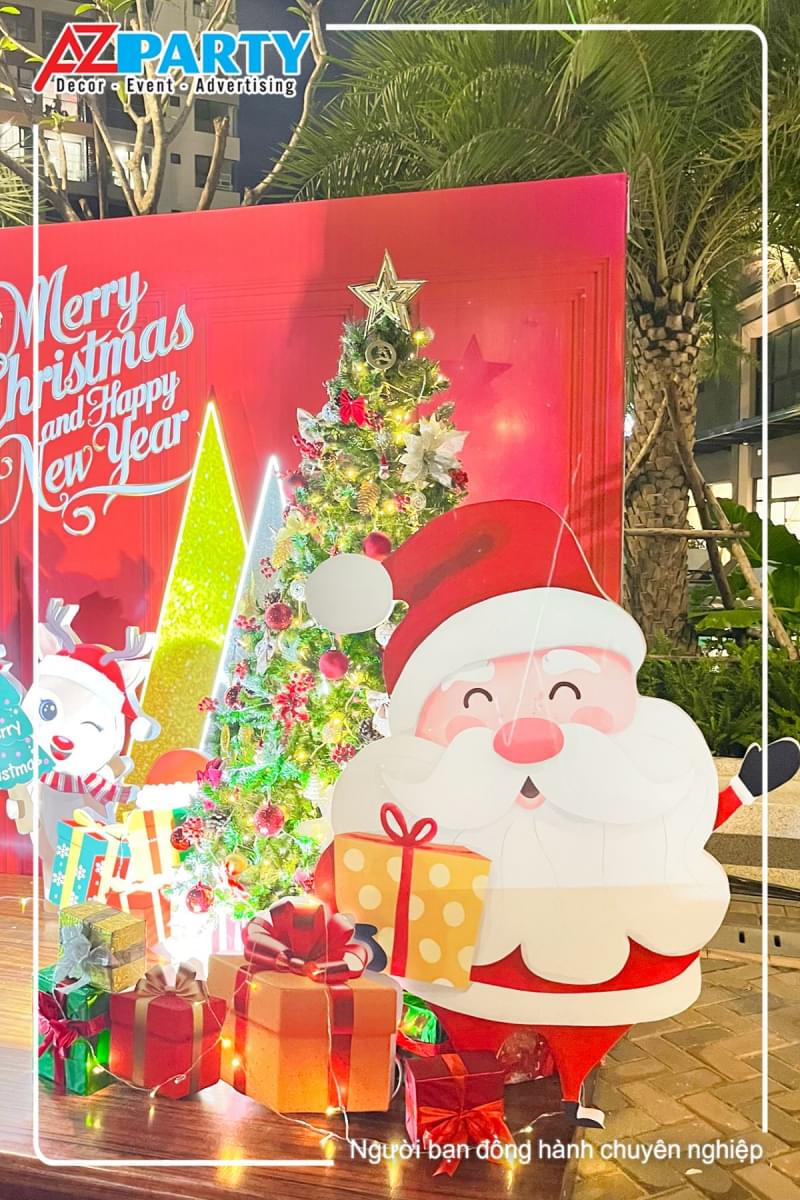 ông già noel tay cầm họp quà, mặc bộ trang phục màu đỏ, đằng sau là cây thông noel lấp lánh ánh đèn vàng